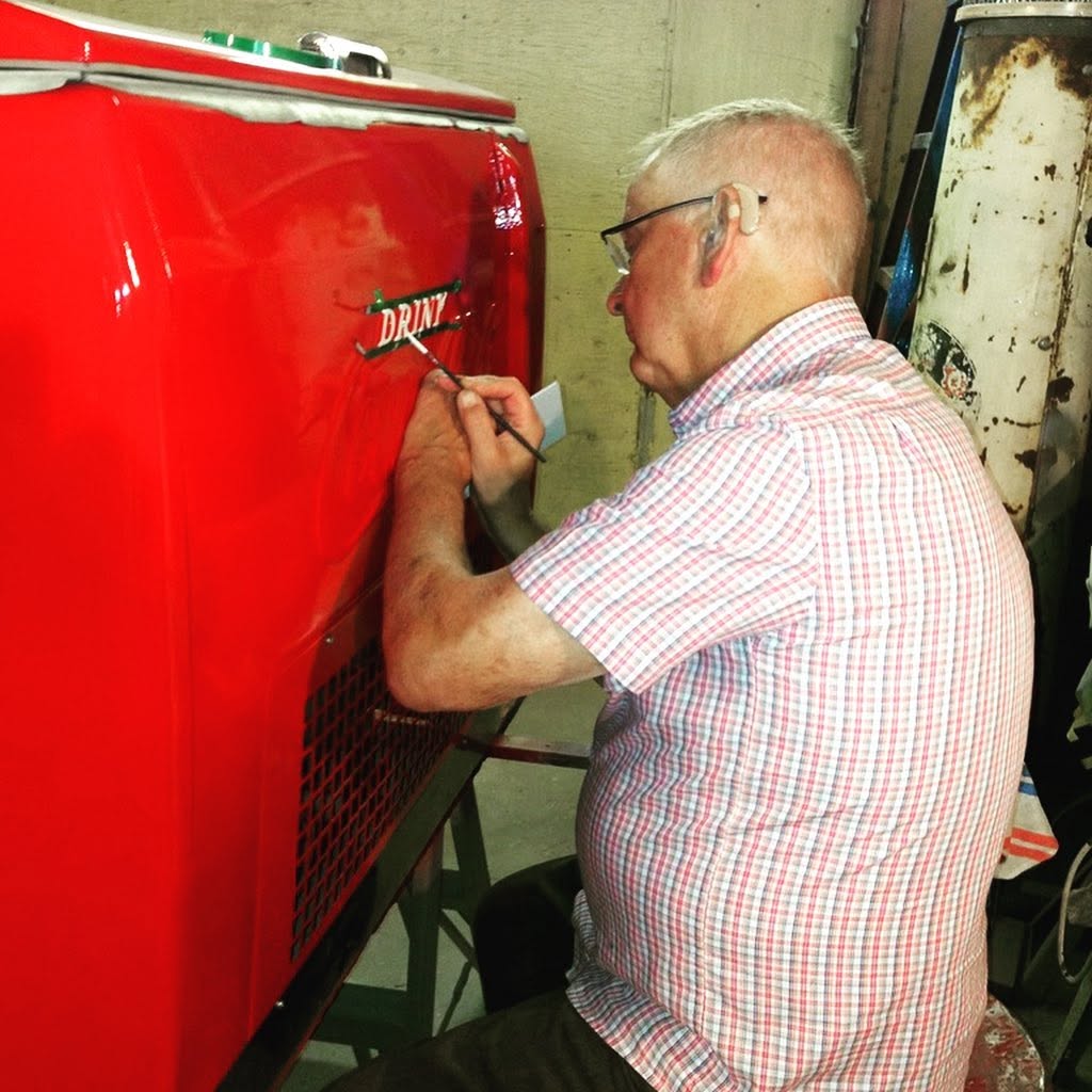 A craftsperson hand-painting red Coca-Cola lettering on a cooler